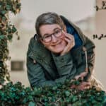 Eine blonde Frau mit kurzen Haaren und Brille die sich auf eine mit Efeu überwucherte Mauer stützt.