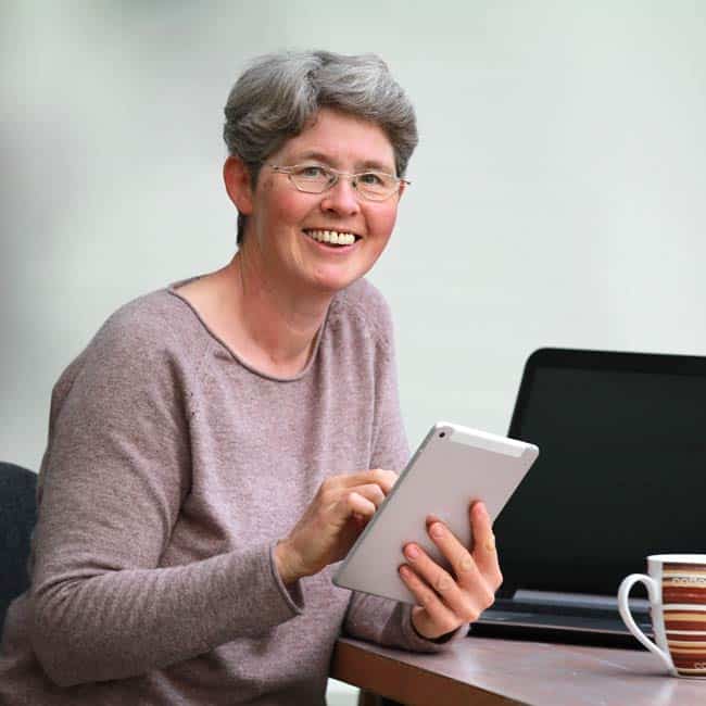 Claudia Barfuss an einem Tisch sitzend, nur der Oberkörper ist sichtbar, mit einem Tablet in der hand und einem beigen Pullover. Im Hintergrund ist ein PC und eine Tasse sichtbar.