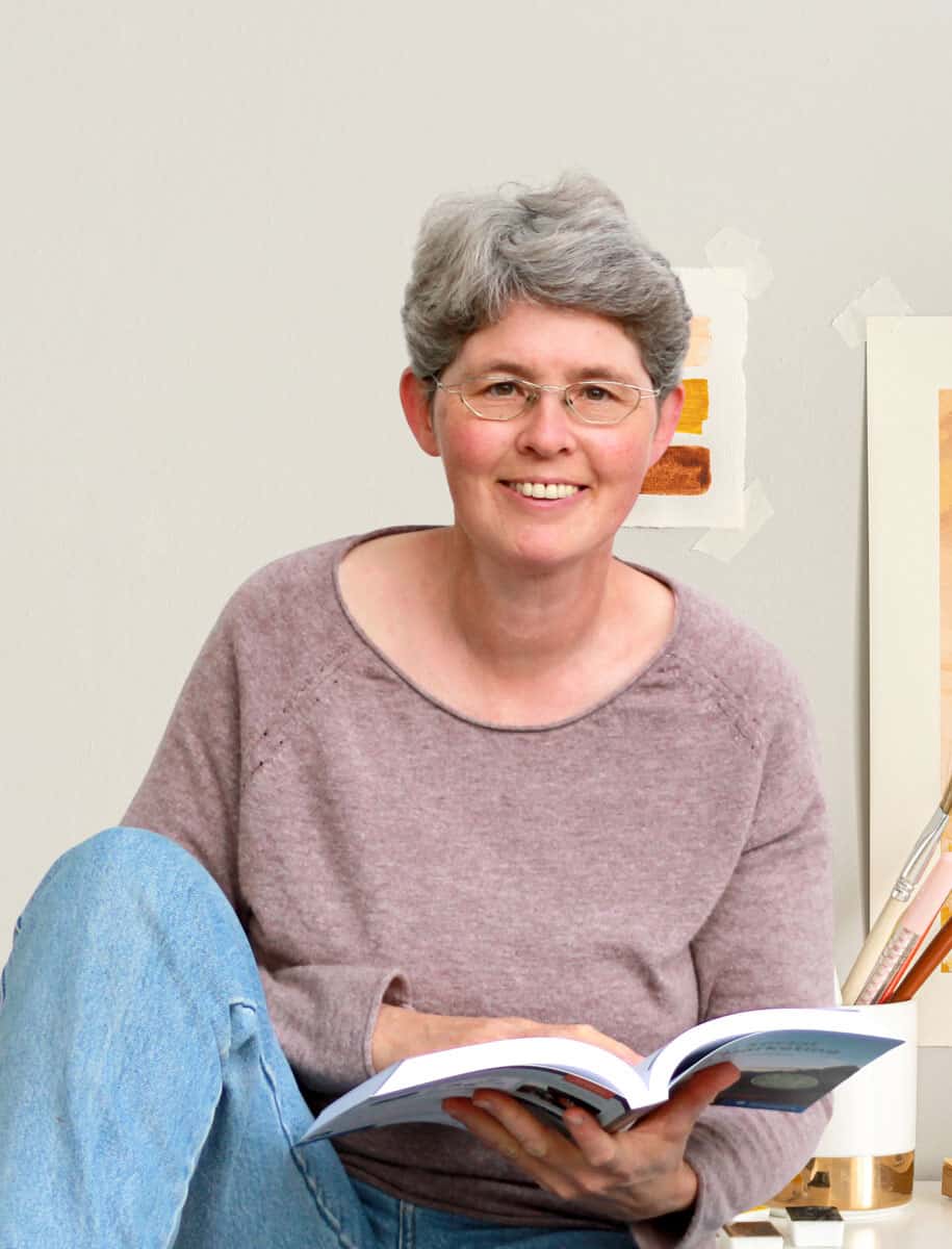 Claudia Barfuss sitzt mit angezogenem rechtem Knie auf einem Tisch und lächelt in die Kamera. In der Hand hält sie ein Buch.