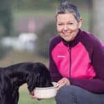 Eine Frau, in einem pinken Oberteil, die kniet und einen schwarzen Hund aus einer Schüssel füttert.