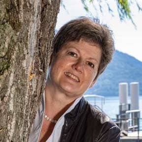 Eine Frau schaut schräg hinter einem Baumstamm hervor, Nur ihr Kopf und ein Teil des Oberkörpers ist zu sehen.